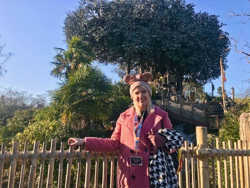Woman in pink coat and Disney parks lanyard standing in front of a fence in the foreground of large trees decorating Adventureland