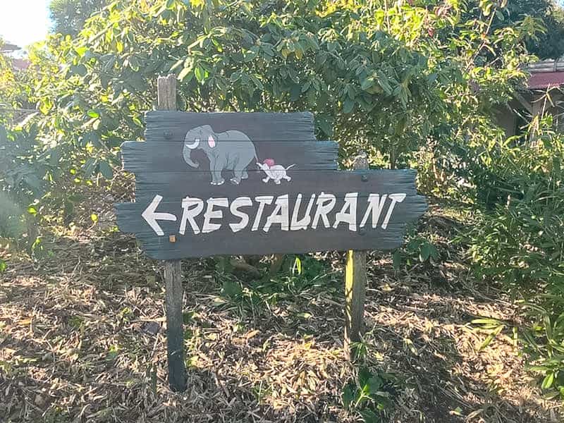 A wooden sign is handpainted with a parent elephant pulling a baby elephant along. The sign reads Restaurant in white paint with an arrow pointing to the left