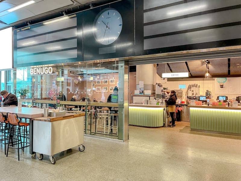 A sandwich shop at St Pancras is brightly lit with a seating area for customers to eat. Above is a grand silver clock