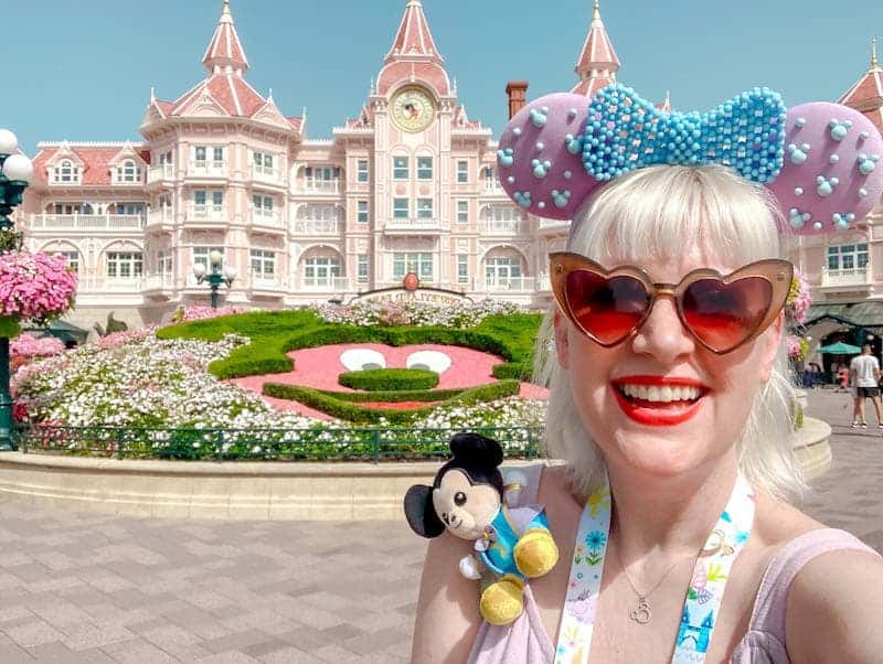 A blonde woman with heart-shaped pink sunglasses and lavendar beaded Mouse Ears is grinning ear to ear in front of the Disneyland Hotel