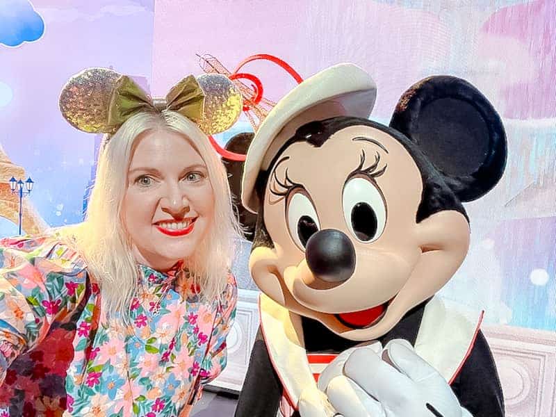 This is a closeup of Minnie Mouse wearing a decorative hat with an Eiffel Tower on it. She is touching heads with a blonde lady wearing a pink floral dress and iridescent mouse ears. Both are smiling sweetly.