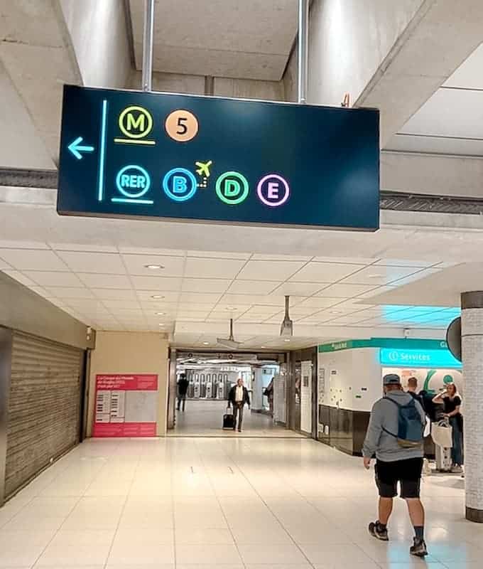 A sign above shows B and D trains, while a man in shorts and a sweatshirt walks underneath it