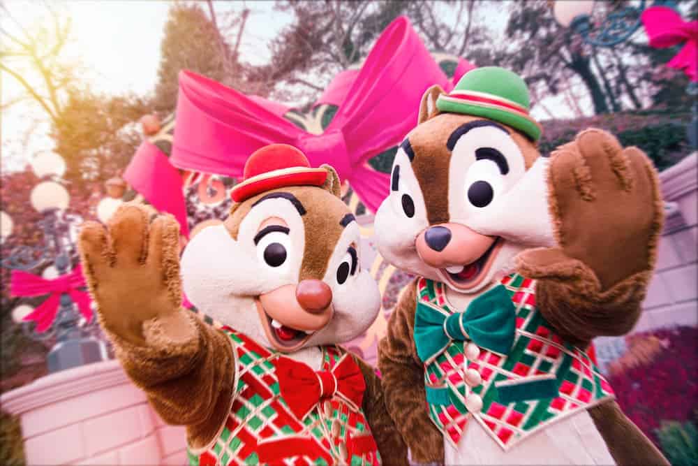 Chip and Dale, two adorable brown chipmunks, are smiling and waving. They are dressed in red and green patterned vests and smart velvet bow ties