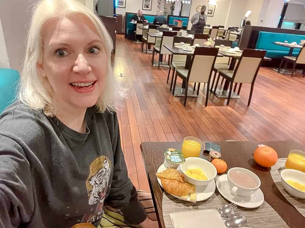 A blonde woman is eating a buffet breakfast of croissant, yoghurt and fruit. There is also hot chocolate. The dining room is modern and clean and there are plenty of tables to choose from