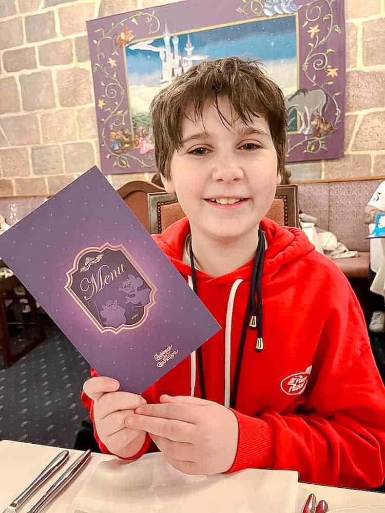 A smiling boy in a red top is holding a purple Auberge de Cendrillon menu for children