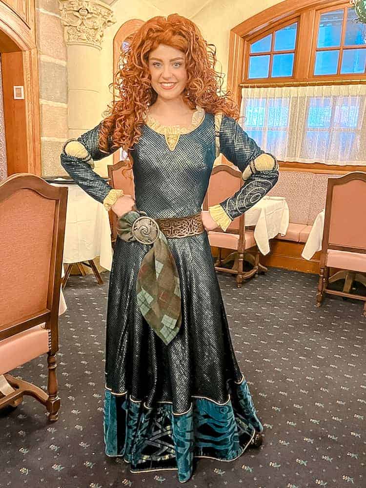 Disney Princess Merida from Brave appears with her tumbling red hair. She stands strong, smiling, in a blue medieval period dress