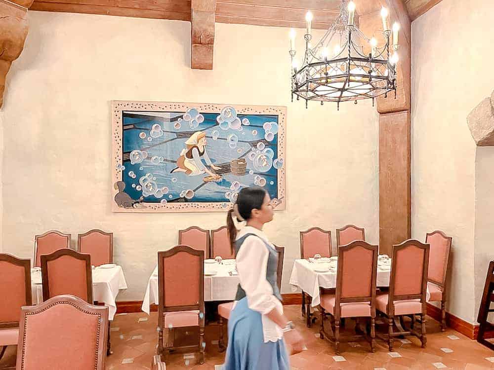 A beautifully themed dining room at Auberge de Cendrillon in Disneyland Paris, featuring elegant pink chairs, a Cinderella mural, and a charming chandelier.