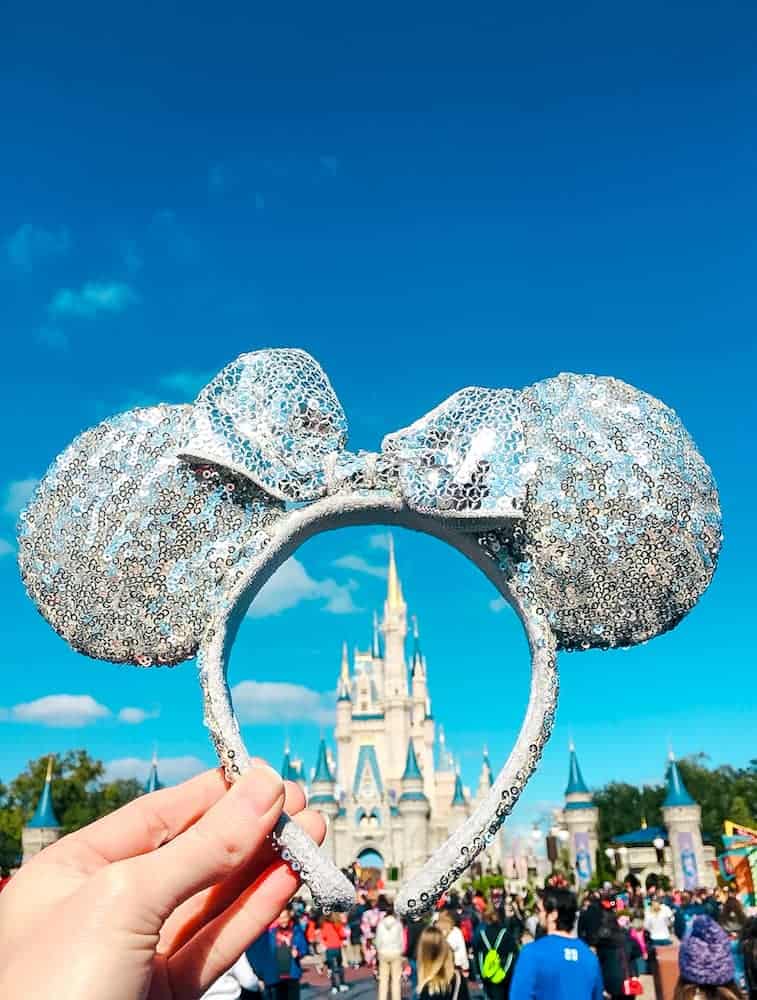 A hand is holding a sparkly Minnie Mouse Ears headband, framing Cinderella's Castle in the distance