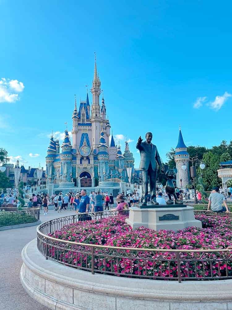 Magic Kingdom's Cinderella Castle is beautiful under a blue sky. In the foreground is Walt Disney's Partners Statue where he is holding Mickey's hand