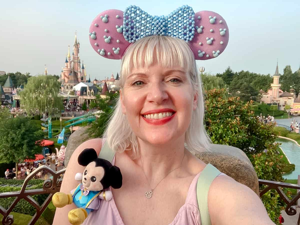 Carrie-Anne Hall blogger of Crepes and Castles - Planning Disneyland Paris for First-timers is in a lavender mouse ears headband and has a Mickey Mouse doll on her shoulder. She is smiling and in the background is a beautiful view of Sleeping Beauty Castle