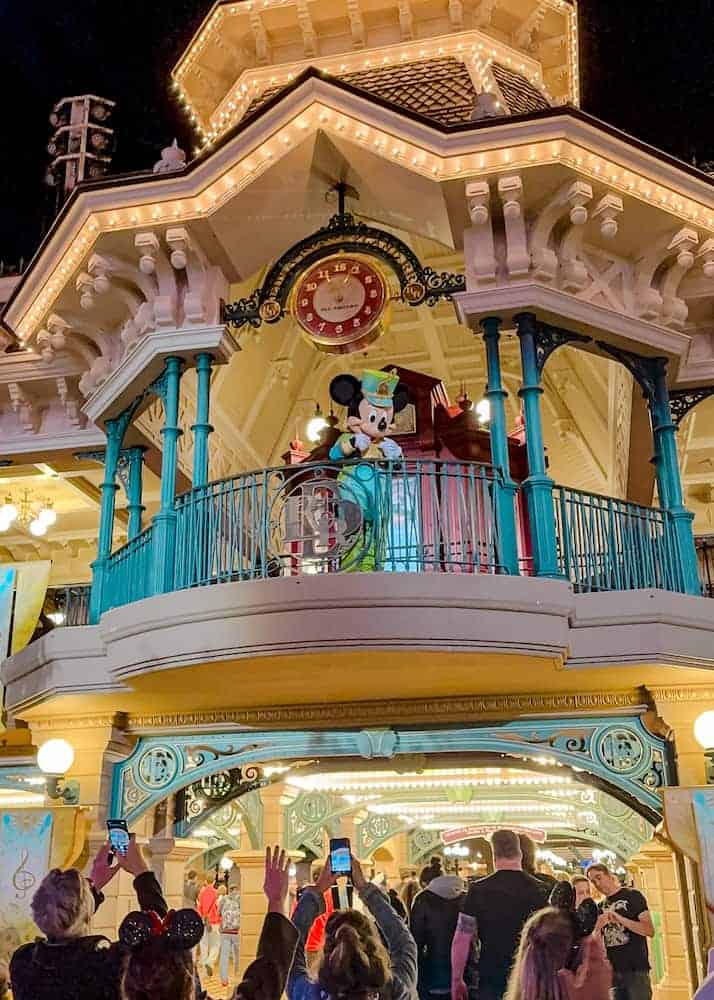Mickey Mouse in a green and gold outfit waving to guests from the Main Street Railway Station balcony at Disneyland Paris, a special evening appearance often seen just after the fireworks — one of the sweetest character moments at the park.