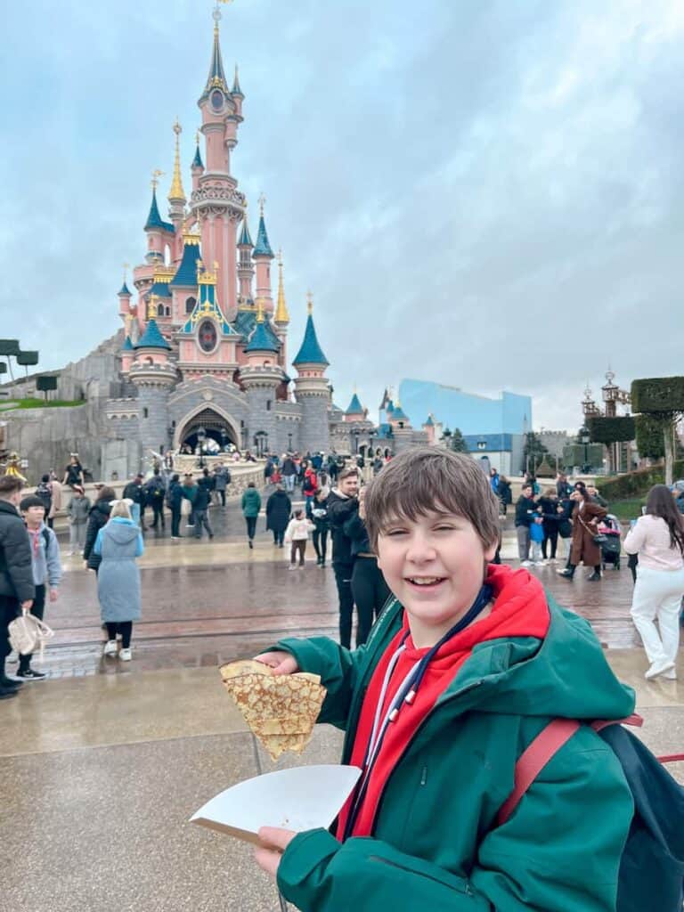 A 13 year old boy is smiling and holding up a Nutella filled crepe in front of Sleeping Beauty Castle in Disneyland Paris