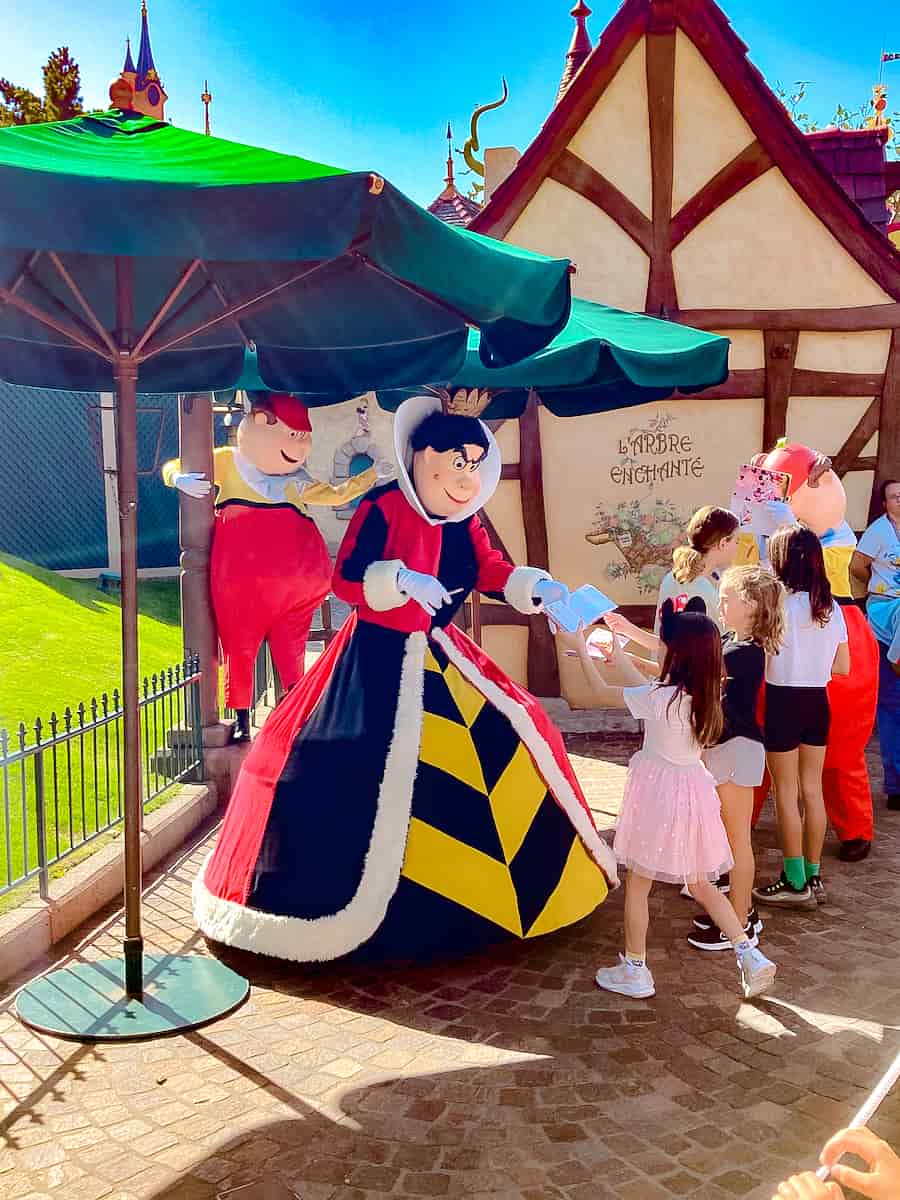 The Queen of Hearts is dressed regally and has a stern face. She is handing a signed autograph book to a little girl.