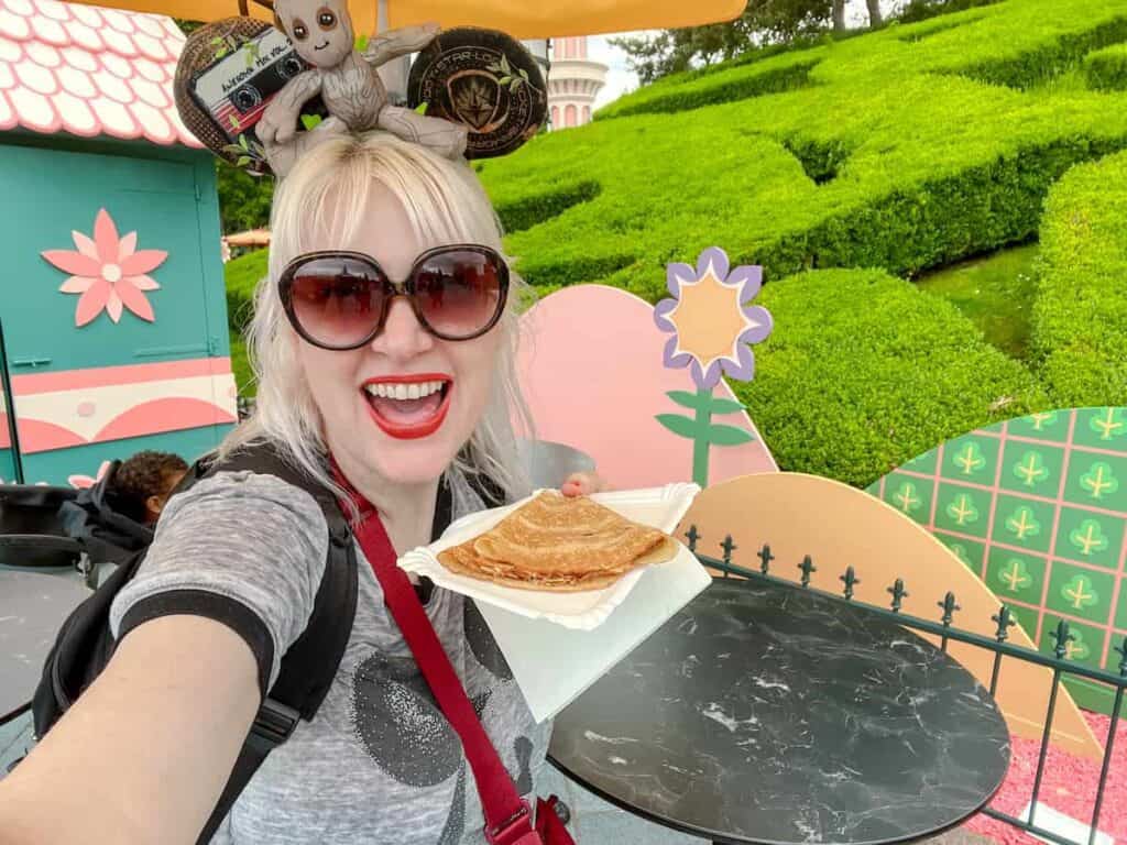 Disneyland Paris blogger Carrie-Anne Hall is holding a yummy plate of crepes. She is perched at a small table in front of a colourful backdrop of it's a small world