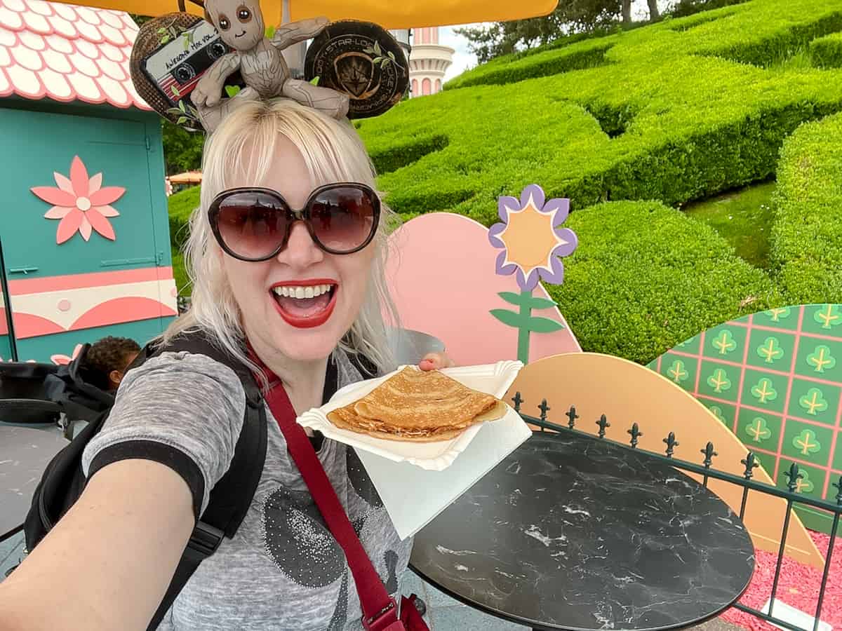 A blonde woman holding a budget-friendly plate of crepes at Disneyland Paris. She has a Groot-themed mouse ears headband and is smiling enthusiastically