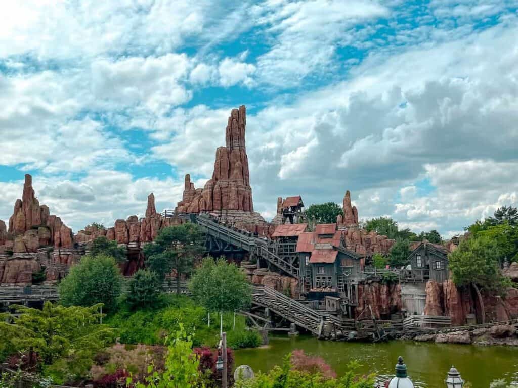 A mine cart chugs up the hill for Big Thunder Mountain while its passengers scream with their hands up. The sky is picture perfect with pillowy clouds rolling by