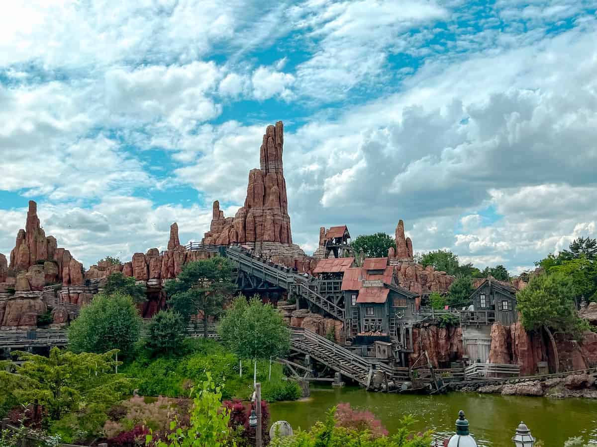 A mine cart chugs up the hill for Big Thunder Mountain while its passengers scream with their hands up. The sky is picture perfect with pillowy clouds rolling by