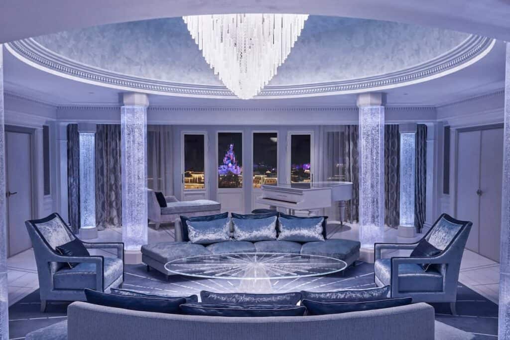 Disneyland Hotel Frozen Royal Suite with ultra-luxurious decor. A piano sits in a pale blue living room with velvet couches. Out the window is a private balcony overlooking the castle at night
