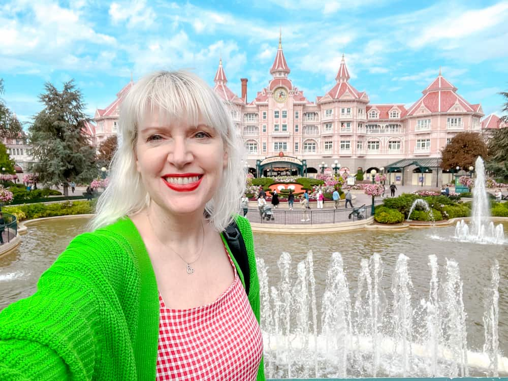 A blonde woman in a red dress and green cardigan is smiling excitedly. Behind her are water fountains and the Disneyland Paris hotel with Club Level Rooms at the top.