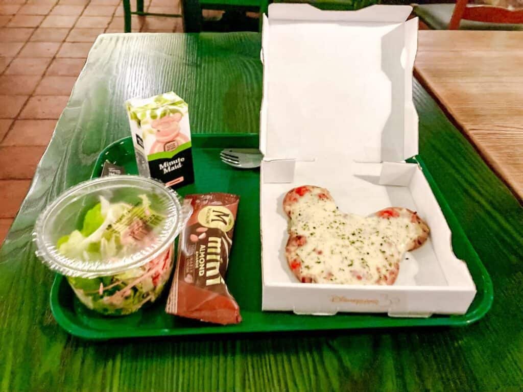 Mickey-shaped pizza, salad, apple juice and mini Magnum on a green tray. This is a kids meal from Colonel Hathis, one of the cheapest ways to eat at Disneyland Paris