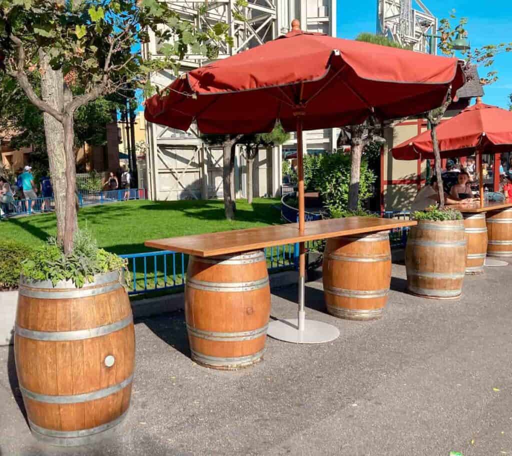 An outdoor bar comprised of barrels serving to prop up a table. An large outdoor umbrella provides shade to Disneyland Paris guests eating their own food in the sunshine