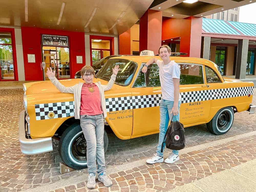 A teen and tween boy in tshirts and jeans are smiling broadly as they pose for a picture in front of a yellow New York Tax cab. Behind them is the door to the Disney Hotel New York: Art of Marvel.