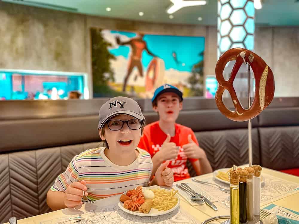 A tween boy in glasses, baseball cap and a striped shirt is smiling exhuberantly, while the teenage boy in the background gives two thumbs up in the science-lab themed Disney restaurant PYM Kitchen.