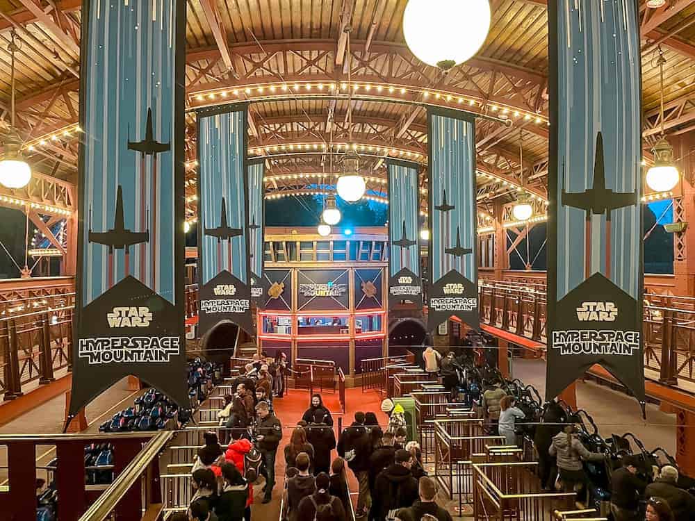 A grand hall filled with Hyperspace Mountain banners. Guests, largely teens and young adults, are gathered in the queues for this high speed roller coaster in Disneyland Paris
