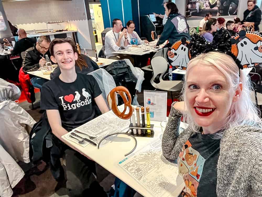 A mum (Disneyland Paris blogger Carrie-Anne Hall) is at a table with her teenage sun inside PYM Kitchen Disneyland Paris. The table has an oversized pretzel and lab bottles for the condiments. Both people are smiling happily