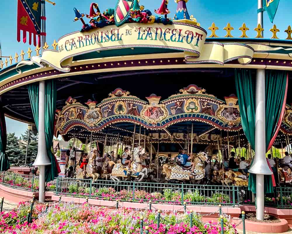 Carrousel de Lancelot in Fantasyland, Disneyland Paris is filled with Disney lovers of all ages from 0 to 99. Toddlers are riding in the carriage, older kids and adults on the moving horses. The vintage carrousel is framed by pretty pink flowers