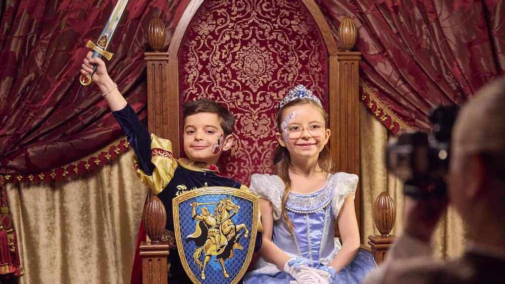 A small boy dressed as a prince raises his sword smiling. Next to him on a throne sits a little girl dressed in facepaint and a princess dress wearing a crown. Both are smiling and enjoying the My Royal Dream Disneyland Paris experience, now open to all guests aged 3 and over.