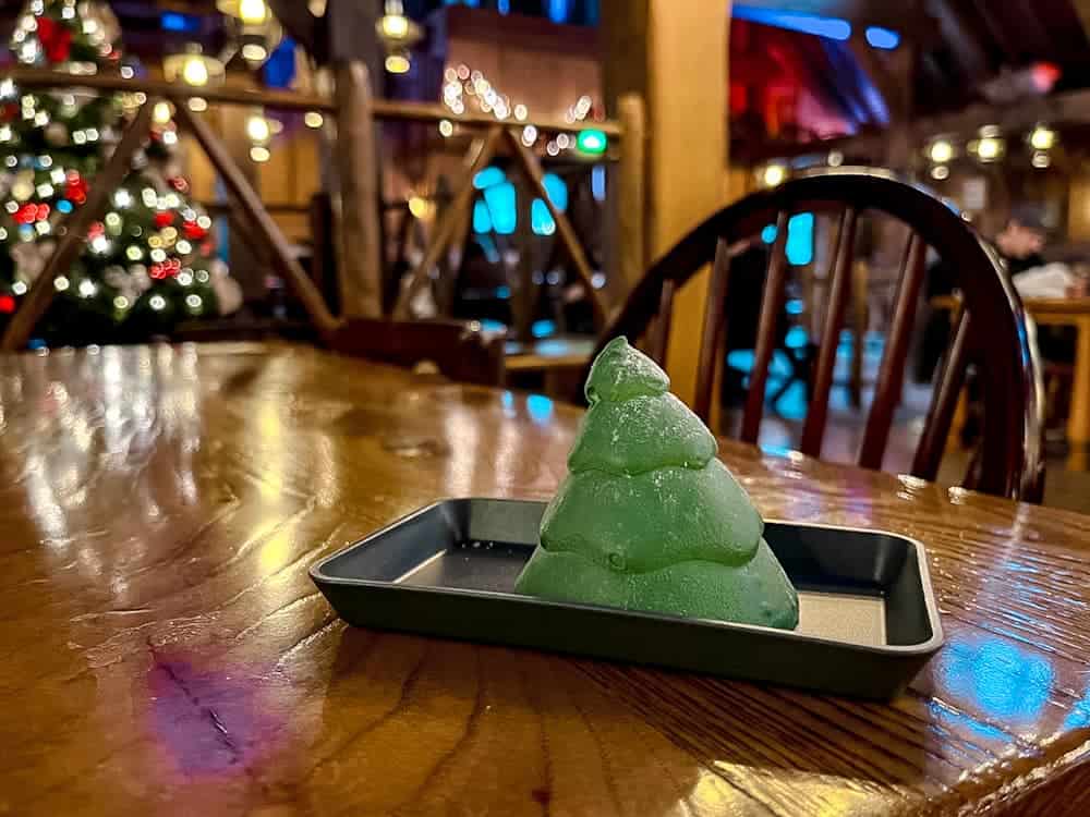 A green Christmas Treet dessert shaped like a Christmas tree, served on a tray inside a warmly lit restaurant at Disneyland Paris, with a decorated Christmas tree glowing in the background.