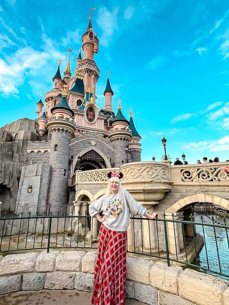A guest (Carrie-Anne Hall) posing in front of Sleeping Beauty Castle at Disneyland Paris, wearing a festive Minnie Mouse sweater and red plaid skirt, against a bright blue sky during the Christmas season.