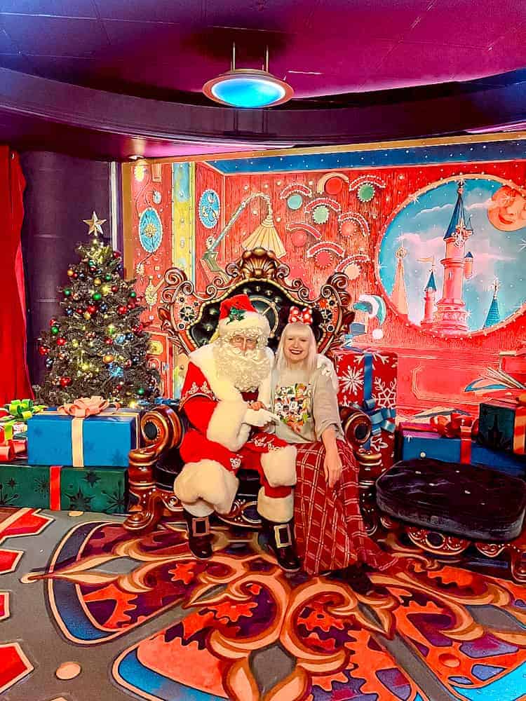 Santa Claus sitting in a festive chair surrounded by Christmas presents and decorations, with a smiling guest (blogger Carrie-Anne Hall) posing beside him in Videopolis at Disneyland Paris. A magical highlight of this Disneyland Paris Christmas review!