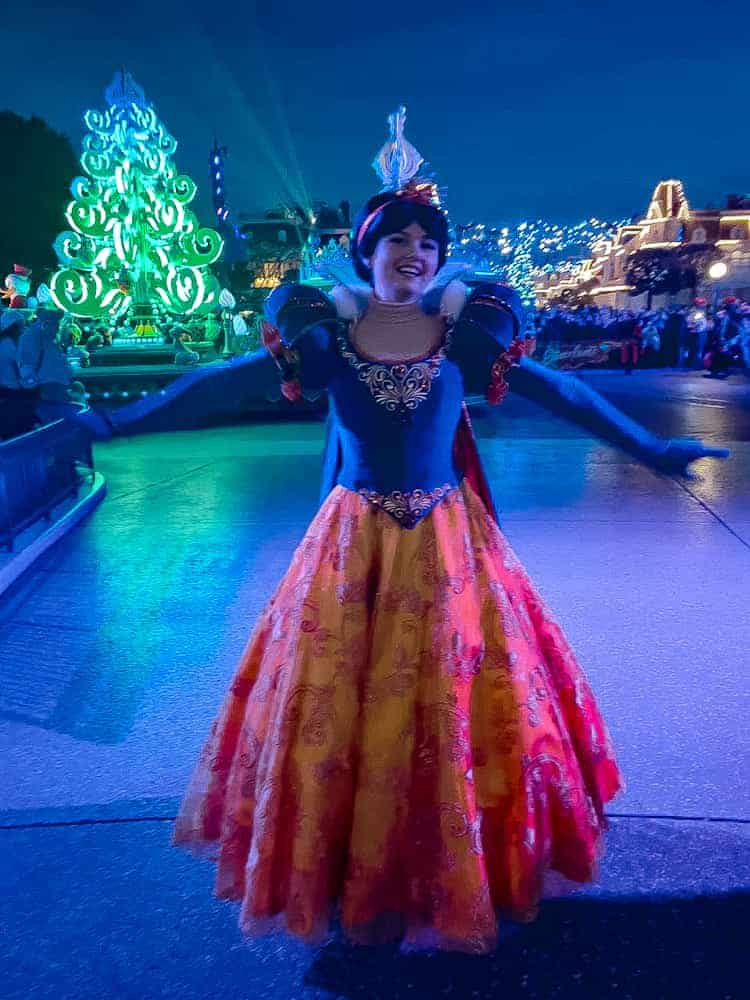 Snow White (from Reserved Access area) gracefully waving during Mickey’s Dazzling Christmas Parade, with the lit-up Christmas tree and dazzling Main Street in the background.