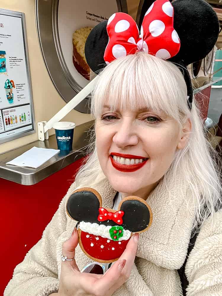 A smiling guest at Disneyland Paris holding a Minnie Mouse-shaped sugar cookie decorated with festive frosting and a holly design. A fun snack moment featured in my Disneyland Paris Christmas review.