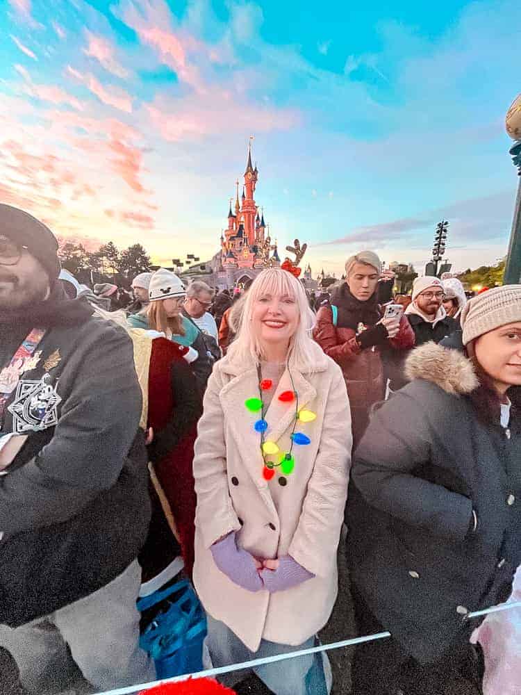 A guest (blogger Carrie-Anne Hall) standing at the front of a parade viewing area - paid Reserved Access - with Sleeping Beauty Castle lit up in the background during Disneyland Paris Christmas festivities.