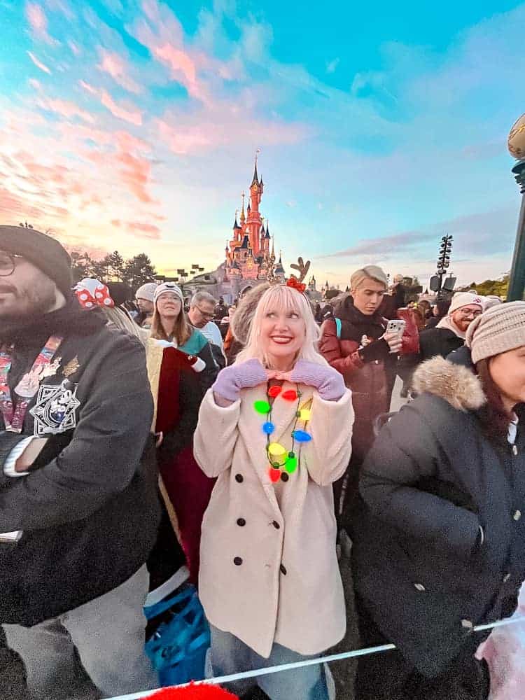 Disneyland Paris blogger Carrie-Anne Hall waits in the Reserved Access Area for Mickey’s Dazzling Christmas Parade, with the Sleeping Beauty Castle glowing against a vibrant sunset in the background.