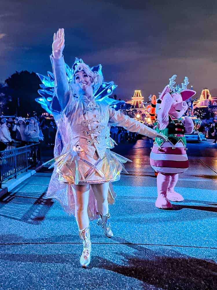 A sparkling performer illuminated in a snowflake outfit waves to the crowd during Mickey’s Dazzling Christmas Parade at Disneyland Paris, with Piglet in a festive costume delighting guests in the background.