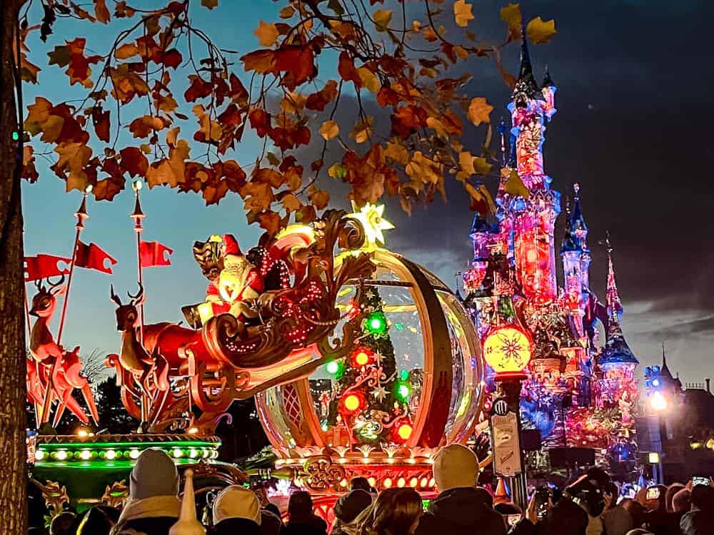 Santa’s sleigh passes in front of the illuminated Sleeping Beauty Castle during Mickey’s Dazzling Christmas Parade at Disneyland Paris, framed by autumn leaves and glowing holiday lights.