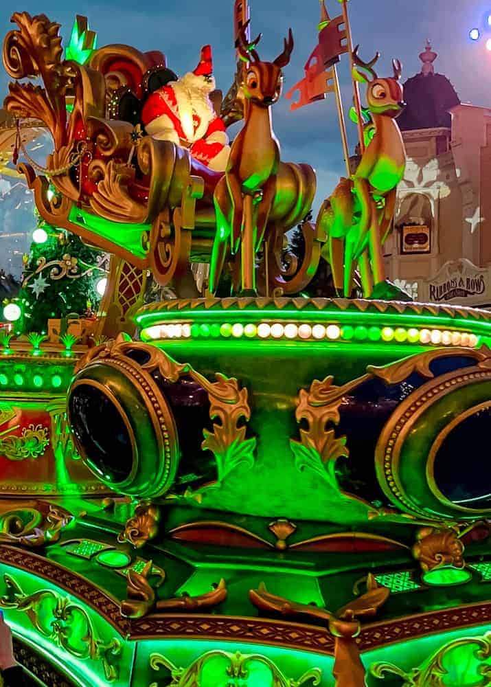 Santa’s sleigh surrounded by glowing reindeer as it passes through the castle hub as an example during Mickey’s Dazzling Christmas Parade: Free vs Paid view experience. This is a free view getting up close to the vibrant green float.