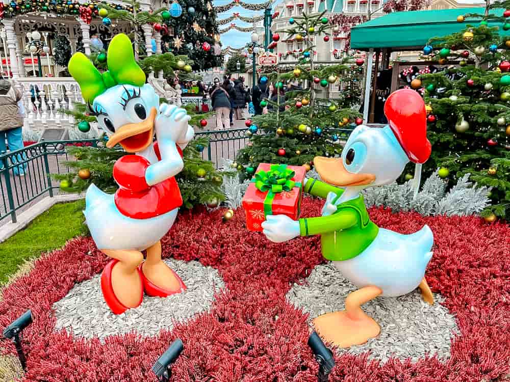 Donald Duck offering Daisy Duck a present in a festive photo spot near Main Street at Disneyland Paris, surrounded by Christmas trees and decorations.