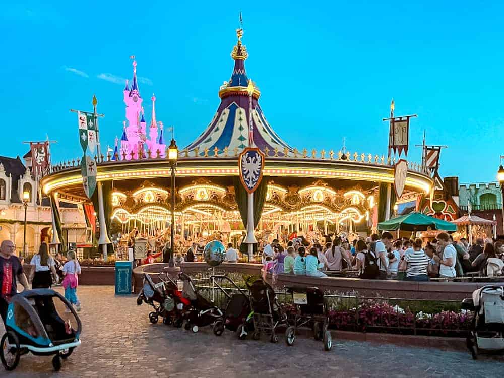 Several strollers and pushchairs are lined up in front of Carrousel de Lancelot in Fantasyland while parents take their children on the vintage carrousel. The attraction is lit in warm yellow light under the twilight sky