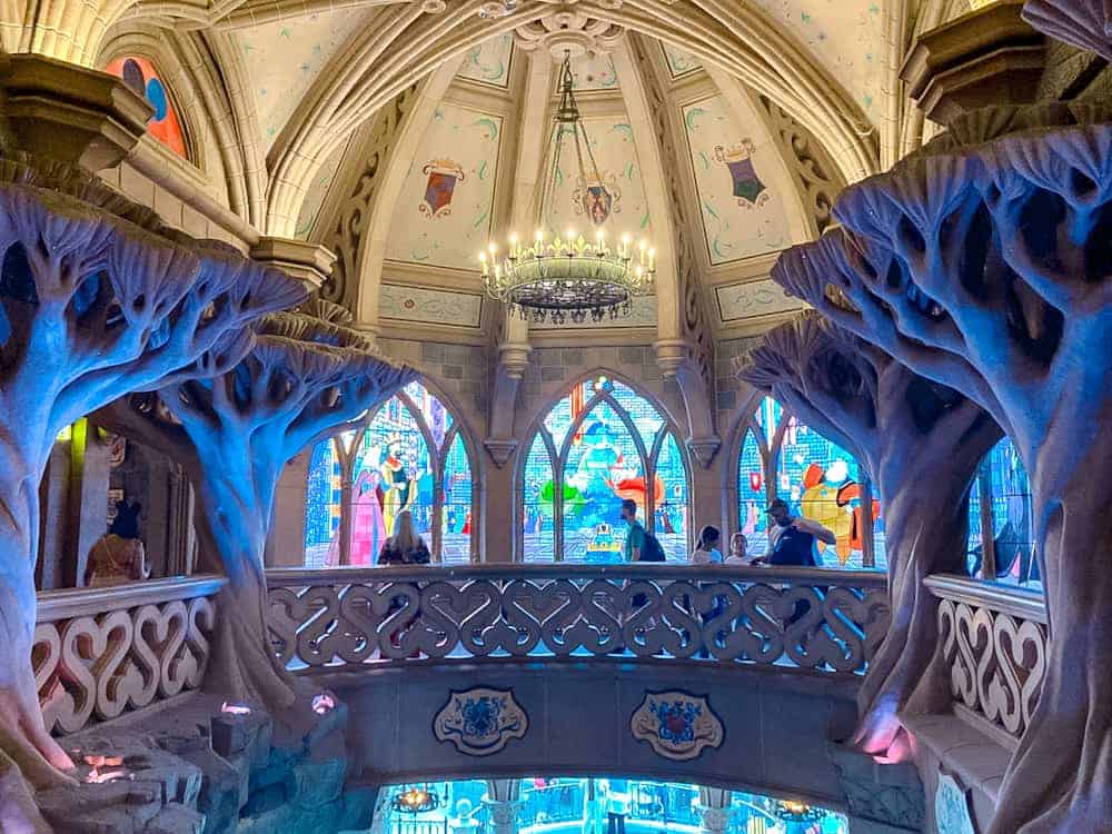 A dad pushes his little one in a stroller through the Sleeping Beauty walkthrough upstairs in a castle. Stained glass windows show scenes from the fairytale while parents enjoy the attraction with their children.