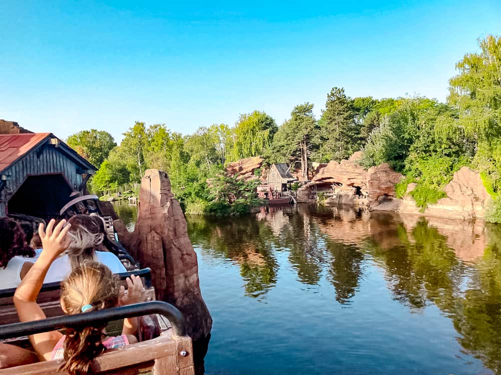 A little girl bravely puts hands up on Big Thunder Mountain, a mine cart themed roller coaster. Beyond is a picturesque lake in Frontierland, one part of the 1-Day Disneyland Paris Itinerary For Families