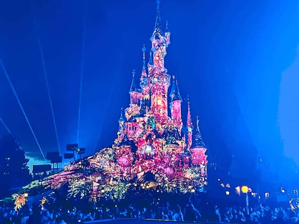 The iconic Sleeping Beauty Castle glowing with vibrant colors and lights against a clear winter sky at Disneyland Paris.
