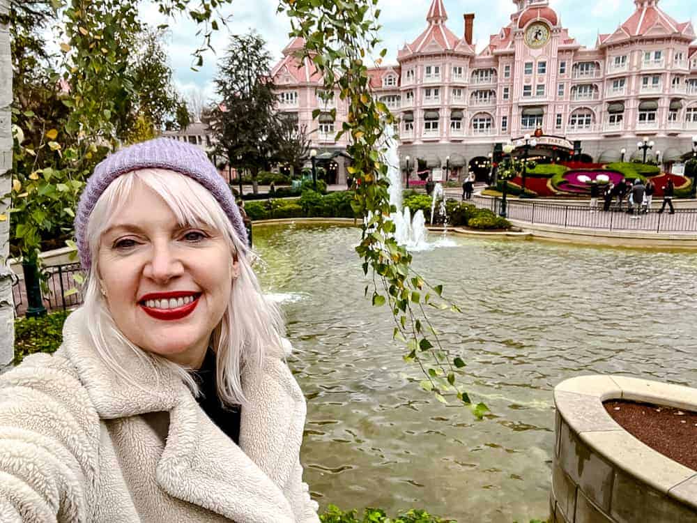 A smiling guest (Disneyland Paris blogger Carrie-Anne Hall) in a lavender beanie posing in front of the iconic Disneyland Paris Hotel on a crisp winter day.