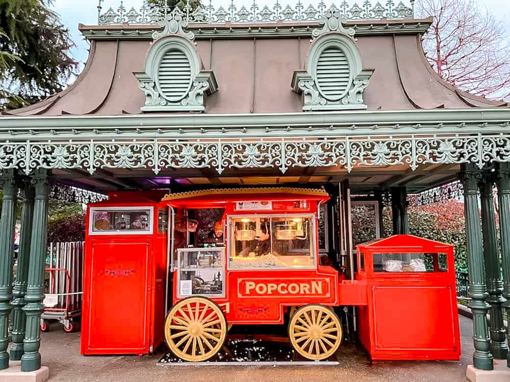 A vibrant red popcorn cart at Disneyland Paris, nestled under ornate ironwork, with popcorn dolls adding to the park’s whimsical storytelling.