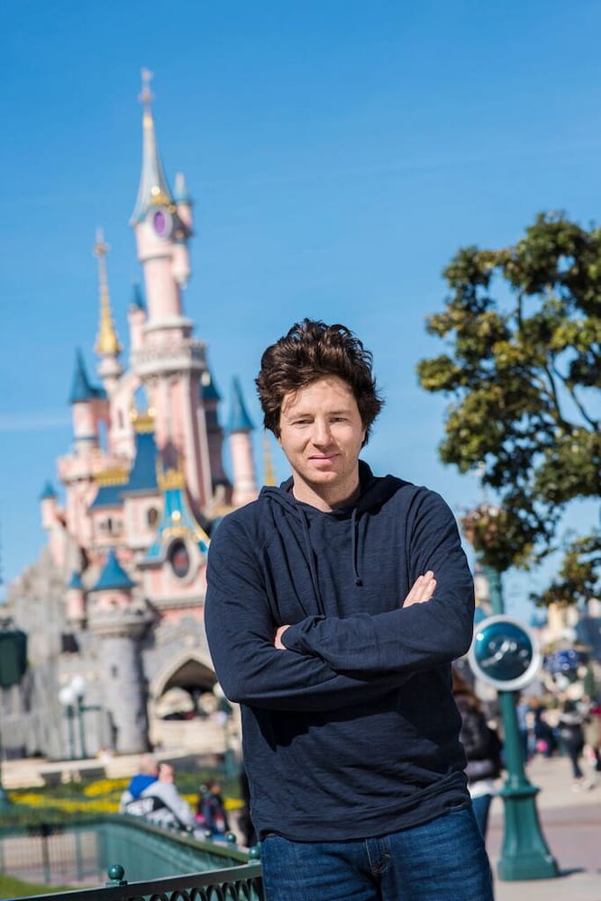 Chef Jean Imbert standing confidently in front of Sleeping Beauty Castle at Disneyland Paris, dressed casually in a navy hoodie on a sunny day. This photo highlights Jean Imbert’s connection to the new Secret Forest Disneyland Paris Restaurant opening in March 2025.