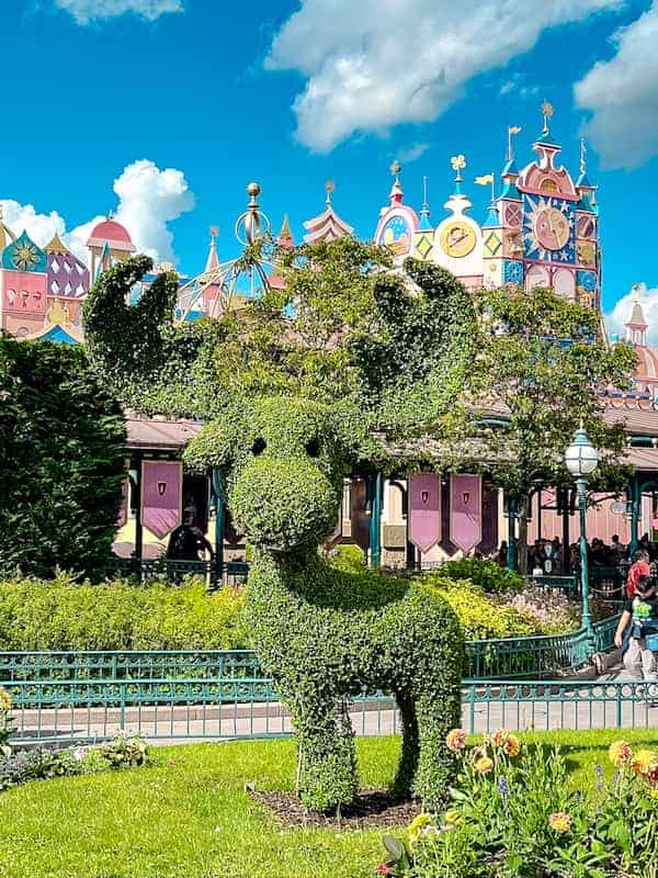 A gorgeous topiary of a reindeer in front of ‘it’s a small world’ at Disneyland Paris in April, surrounded by vibrant spring flowers and lush greenery.